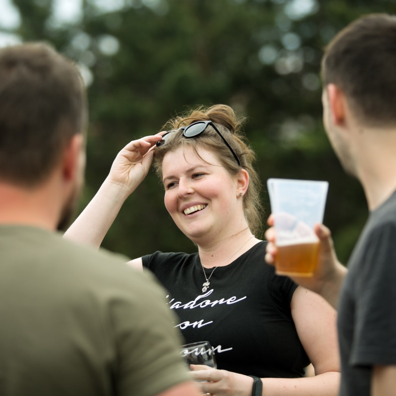 Žena s úsměvem mezi kolegy na venkovním firemním setkání. Drží si sluneční brýle, kolem ní lidé s plastovými kelímky a pivem.