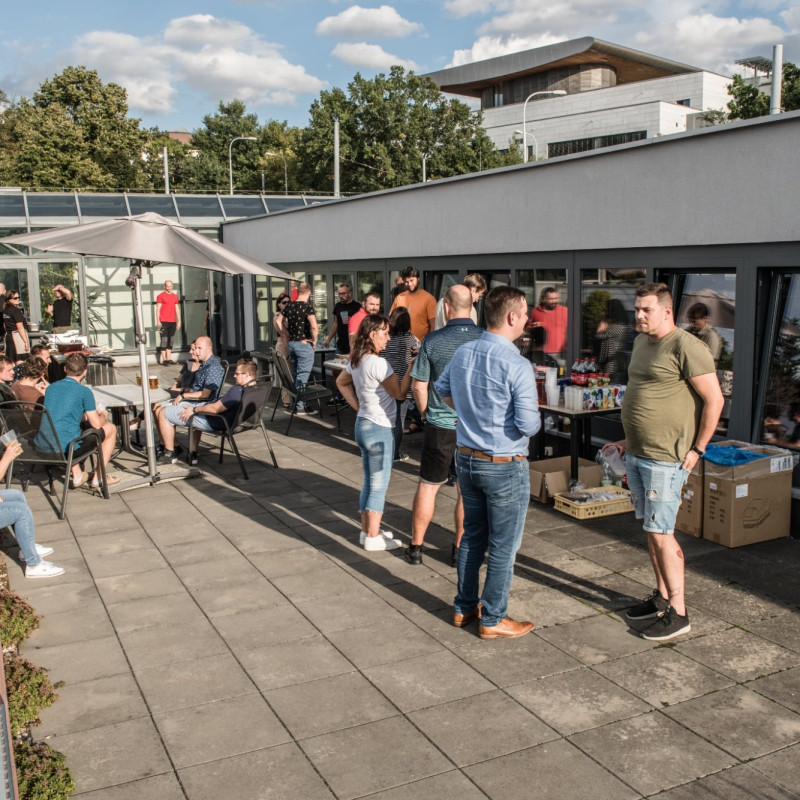 Lidé se setkávají na střešní terase firmy při grilovací akci, povídají si u stolů nebo ve skupinkách, v pozadí je prosklená budova a zeleň.